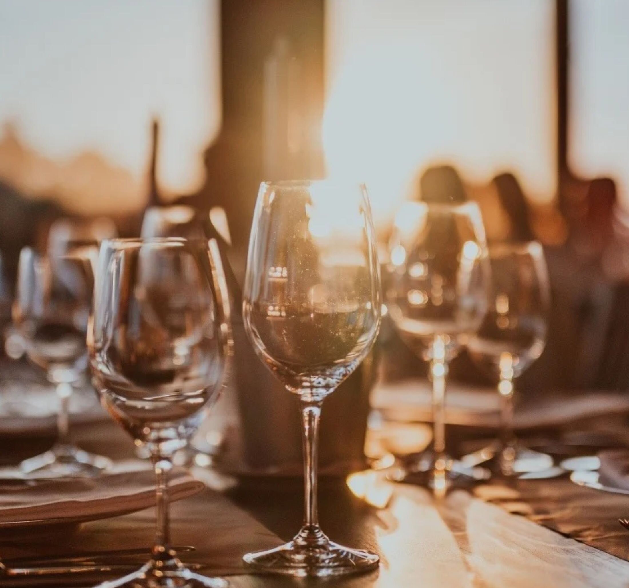 golden hour sunlight filters through empty wine glasses in a fine dining room where a table is set with wine glasses and people sit around tables in the background