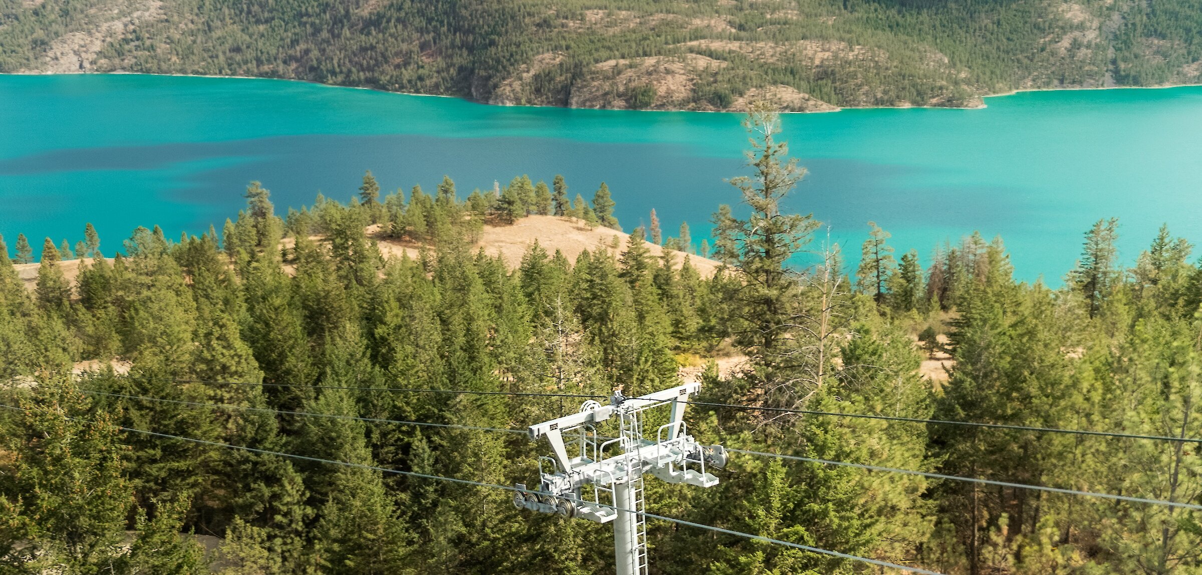 Aerial view of a newly installed steel gondola lift tower surrounded by pine forest overlooking the turquoise waters of Kalamalka Lake at the Okanagan Gondola