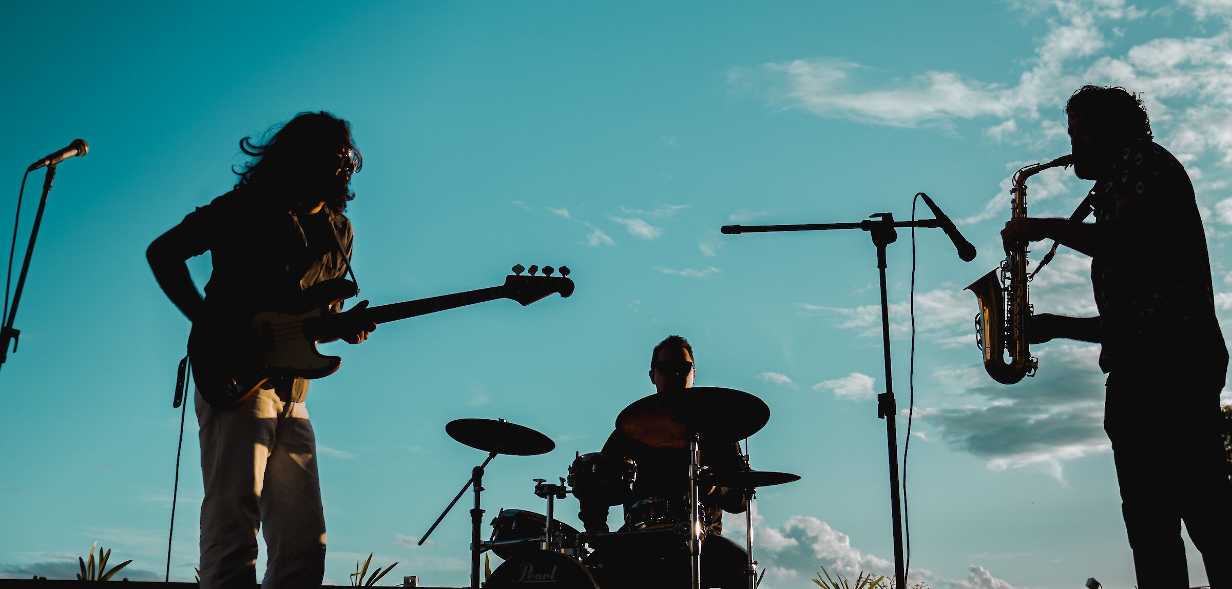 Silhouette of a live band with a guitarist, drummer, and saxophonist performing outdoors against a bright blue sky