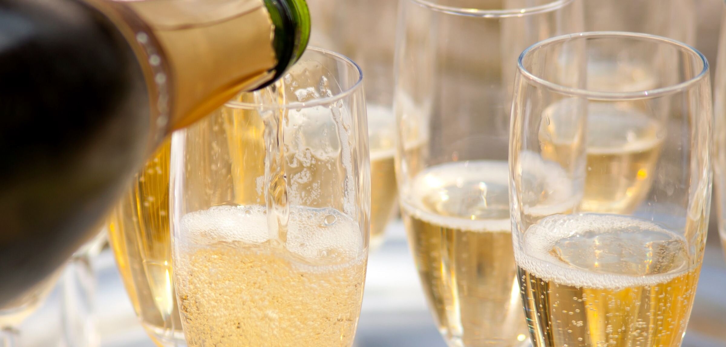 Close-up of champagne being poured from a bottle into a clear champagne flute with additional glasses in the background..