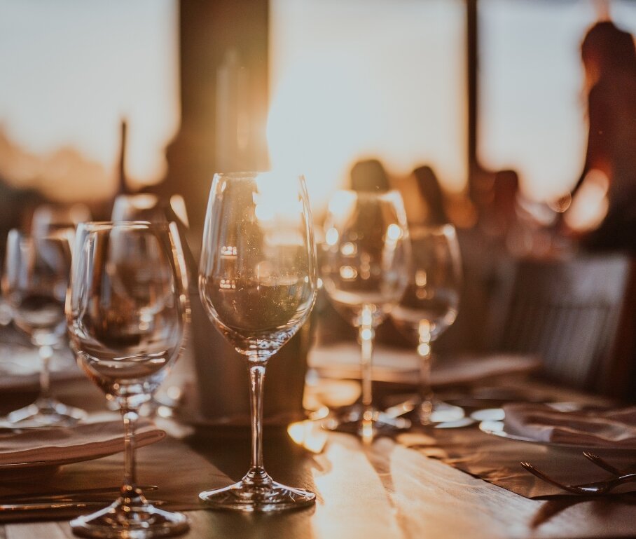 golden hour sunlight filters into a fine dining room where a table is set with wine glasses and a woman stands over a table in the background
