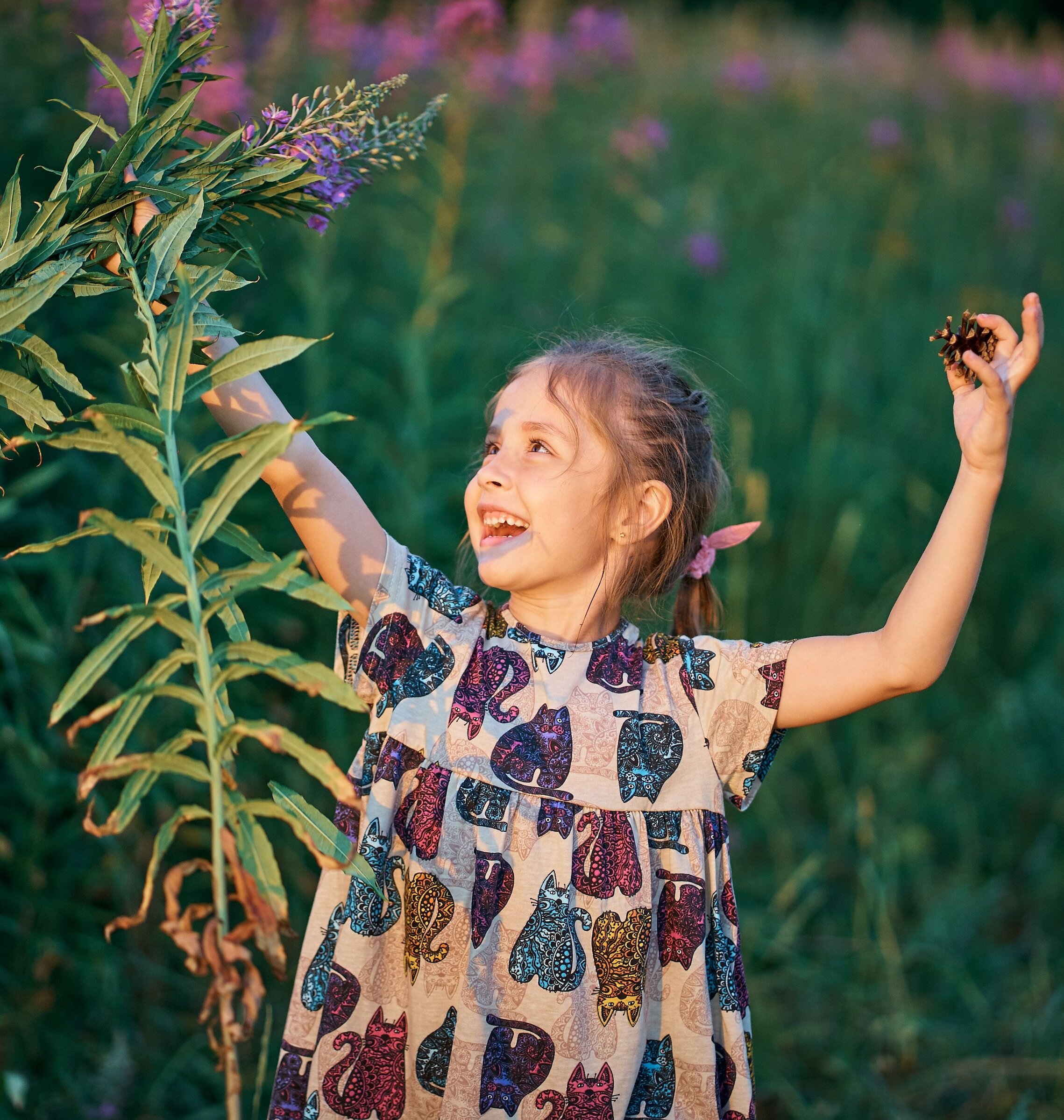 Happy young girl holding a pinecone and playing near tall purple wildflowers along a mountain nature trail