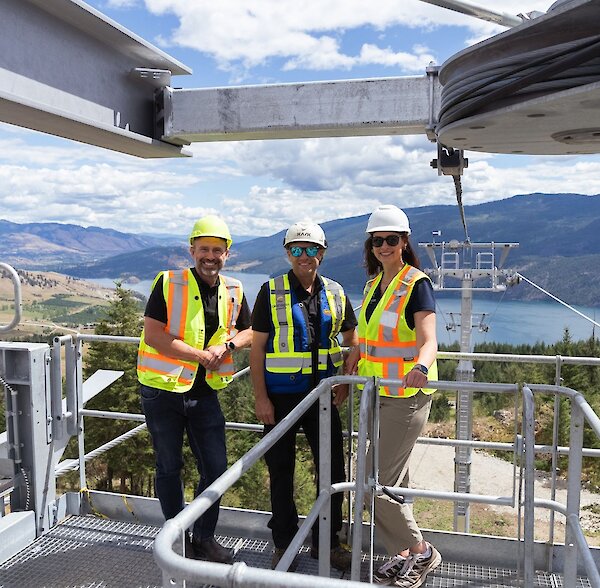 Group photo of three people standing on gondola terminal overlooking Kalamalka Lake with mountains in the background