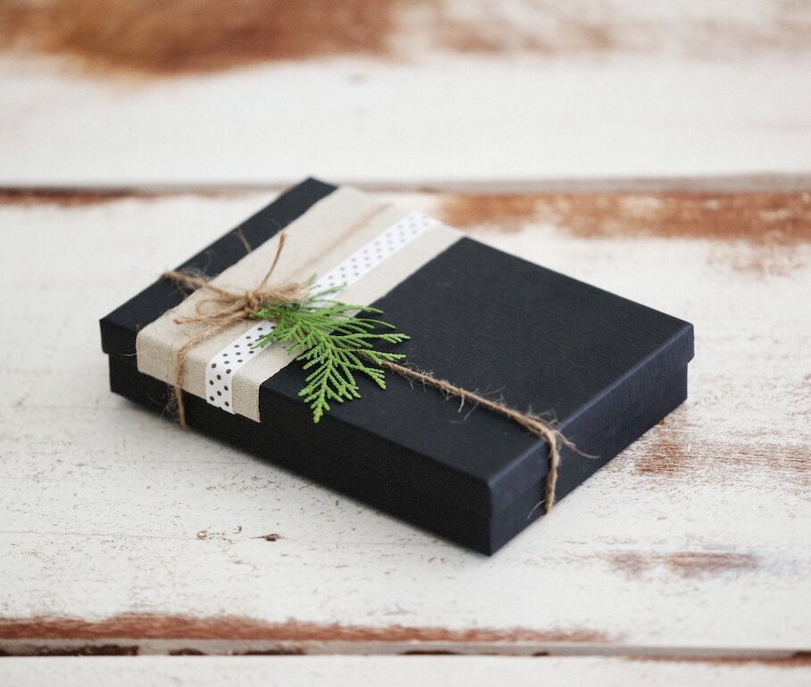 Wrapped black gift box with twine ribbon and evergreen sprig on a rustic white wooden surface